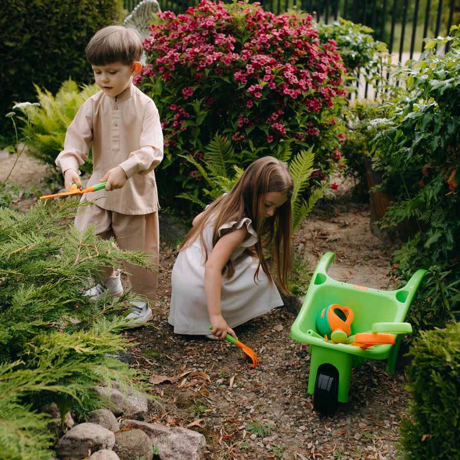 Deux enfants jardinent avec une brouette dans un jardin fleuri.